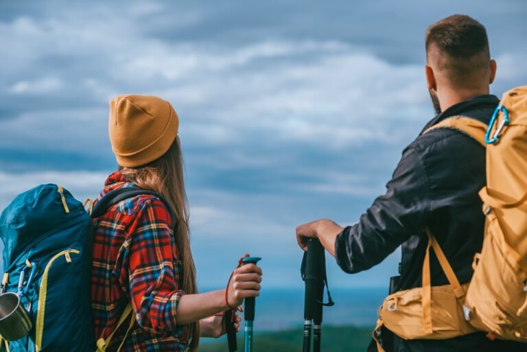 Two hikers with backpacks and trekking poles pausing on a coastal trail under a cloudy sky