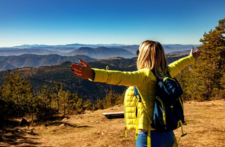 Hiker in a yellow jacket with a backpack standing with open arms in front of a mountain range