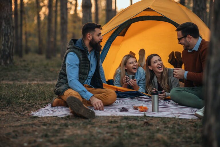 Four friends sitting on a blanket outside an orange dome tent in a pine forest