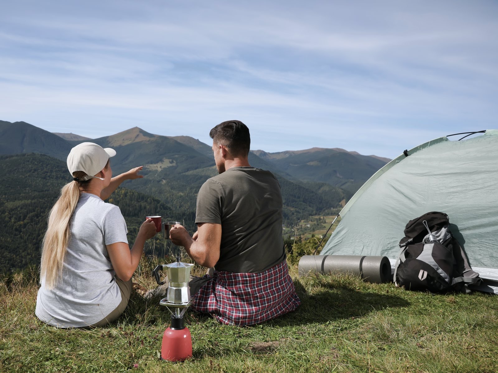 Two campers drinking coffee beside their tent, looking out over green mountain ridges