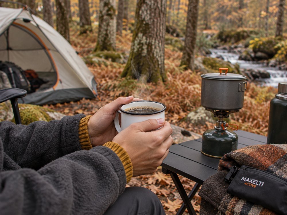 Hands holding an enamel mug of coffee beside a camp stove with a tent and pine trunks behind