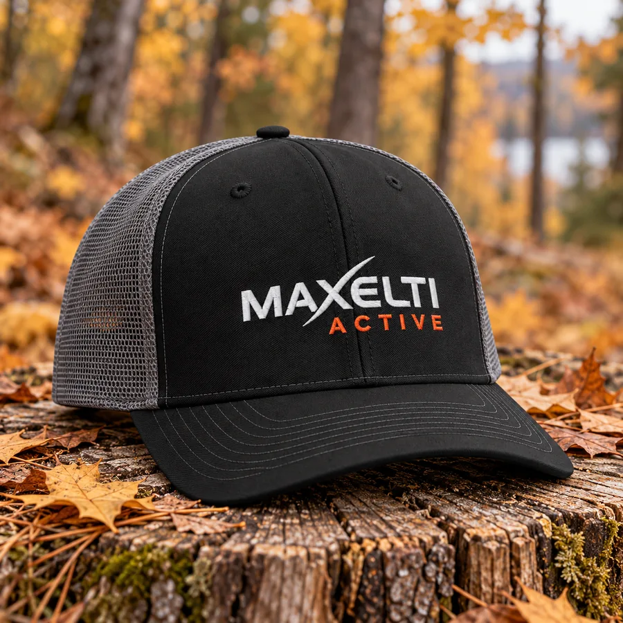 Black and charcoal mesh trucker cap with the MAXELTI Active logo resting on a log stump in an autumn forest clearing